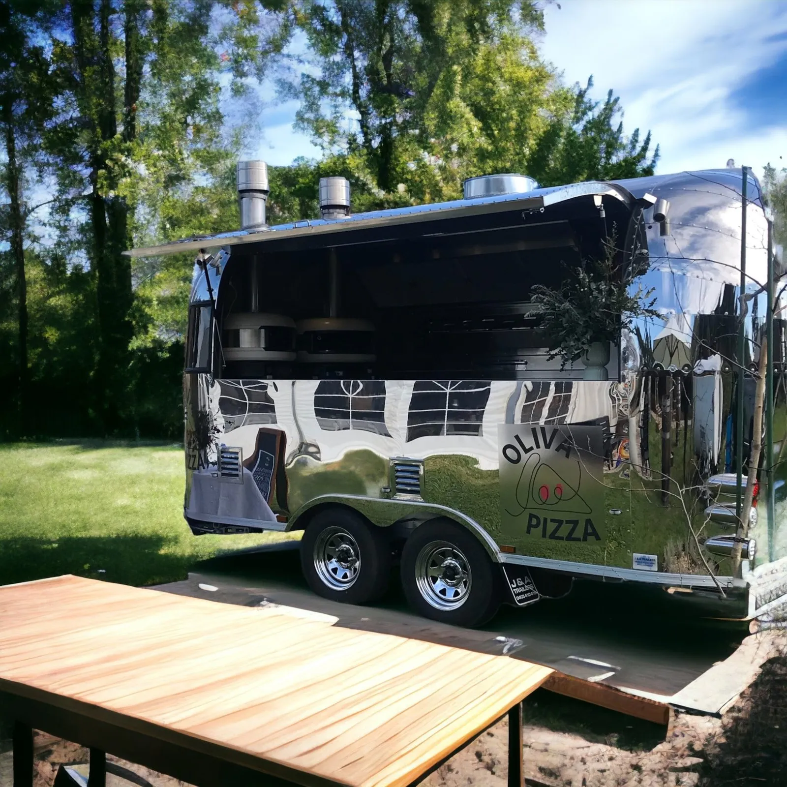 The chrome trailer on a lawn beside a long timber table, trees behind